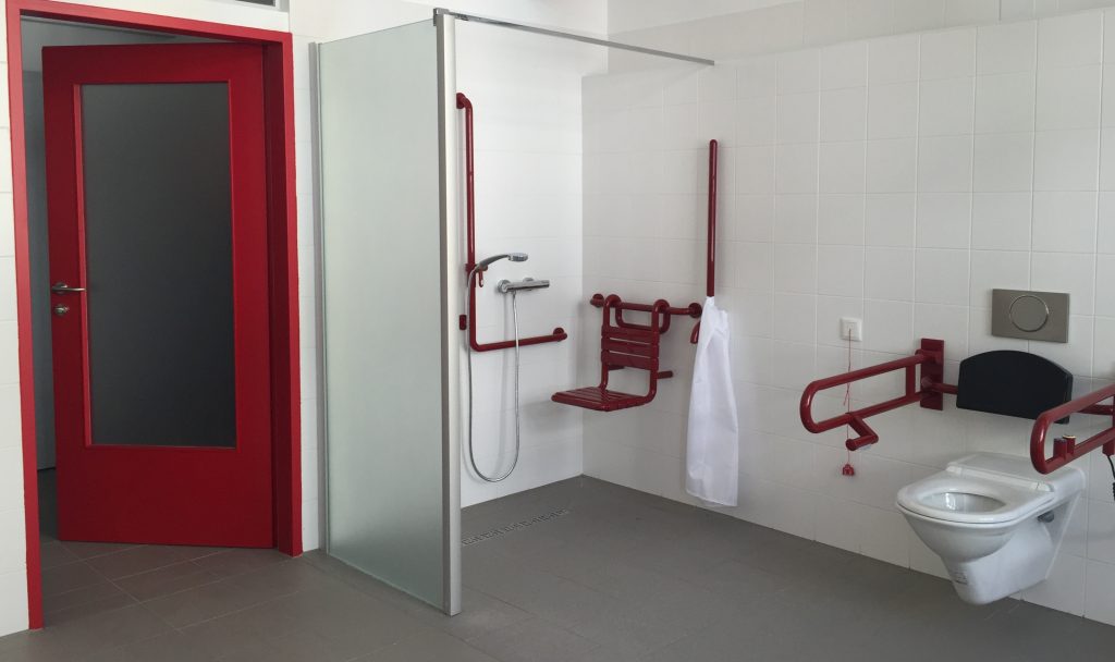 Foto Sanitärraum mit barrierefreier Dusche und WC, dunkelgrauem Boden, weißen Wänden und roten Kontrastelementen