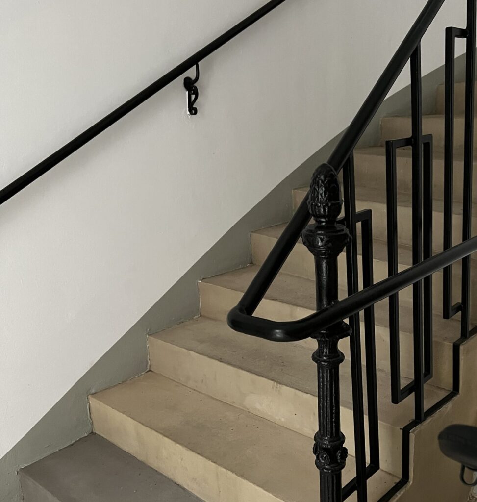 Foto einer Treppe mit sandsteinfarbenen geradkantigen Stufen und schwarzem Handlauf und Geländer. 